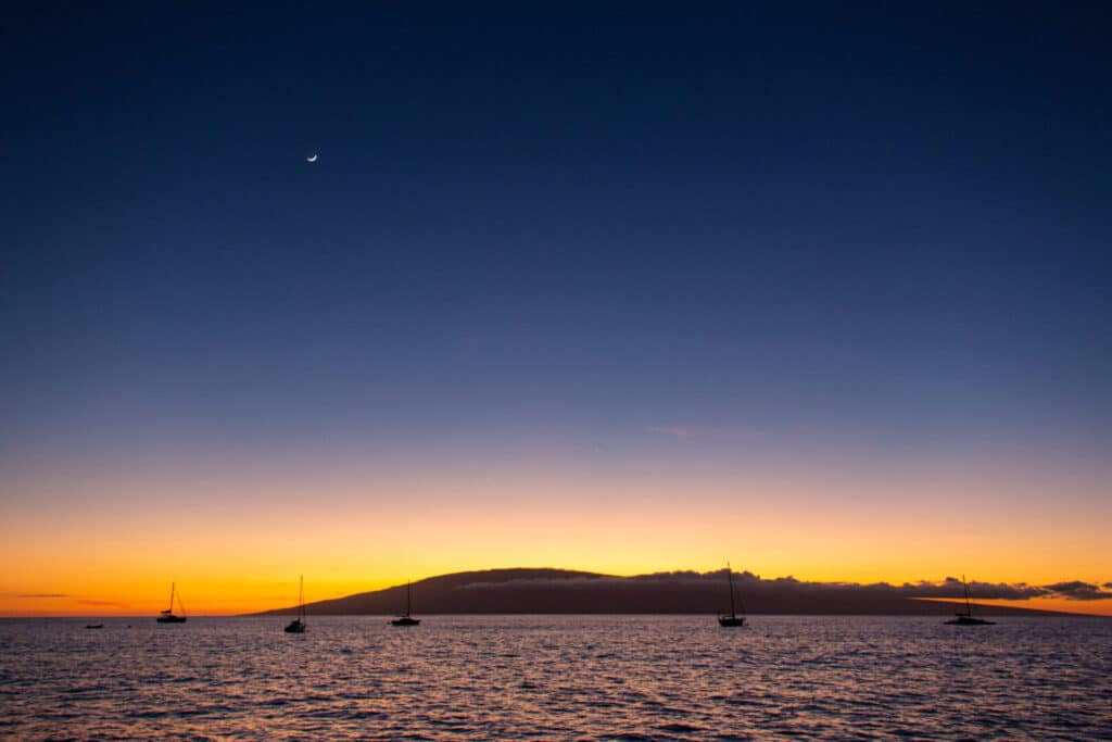 Sunset over a calm sea with silhouetted boats and hills.