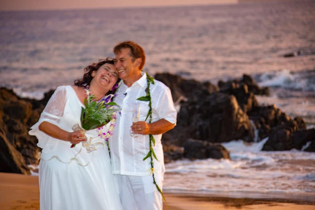 A couple in white attire embraces on a beach during sunset.