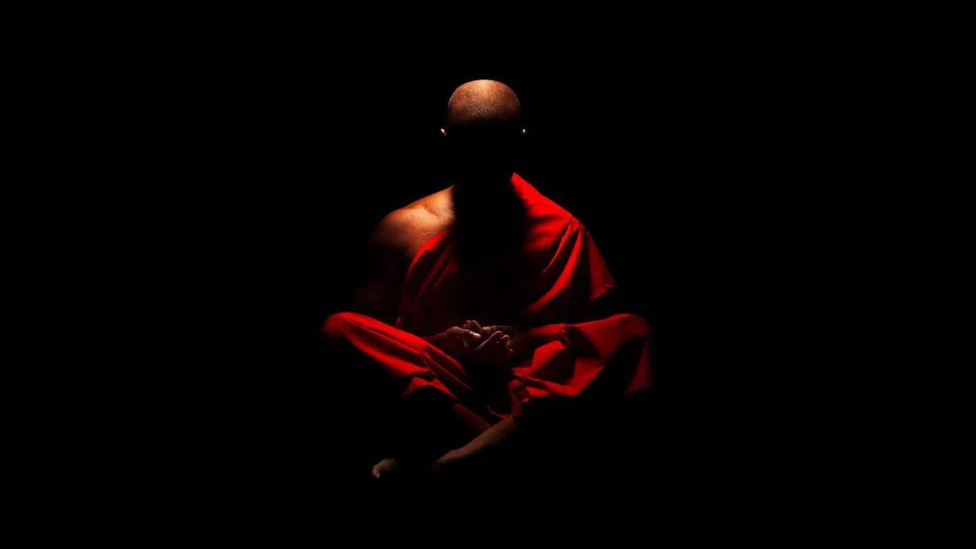 A monk meditating in darkness with red robes illuminated.