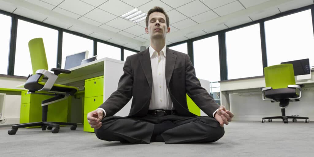 A man in a suit meditating cross-legged in an office.
