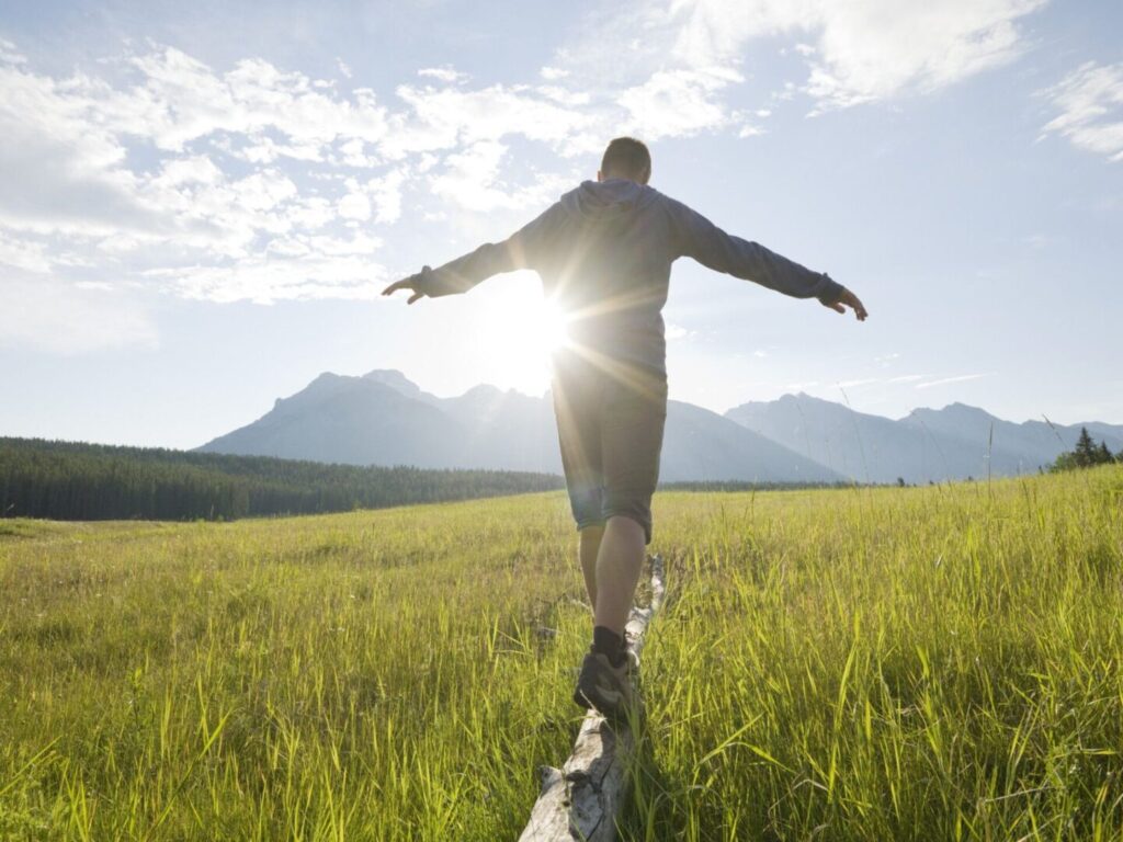 Person balancing on a narrow path in a grassy field with mountains in the background.