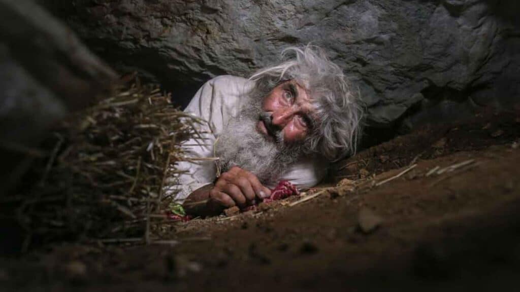 A man with a beard peeking out from a dark cave.