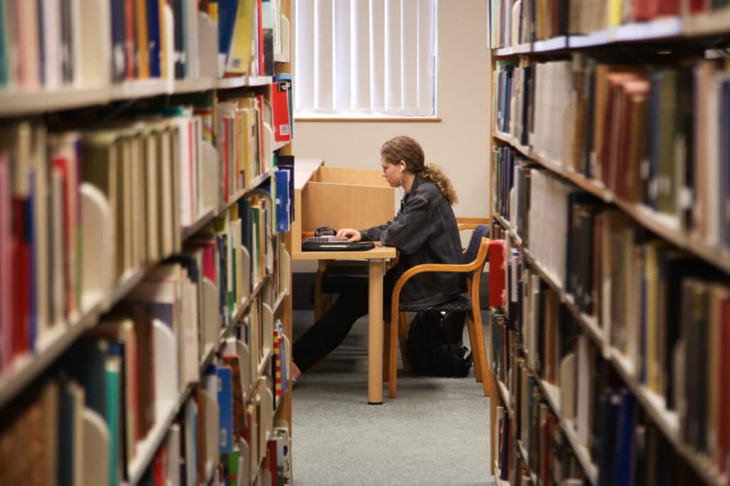 A person studying at a desk in a library aisle.