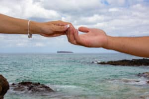 Two hands reaching out over the ocean under a cloudy sky.