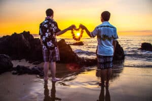 Two people at sunset forming a heart with their hands by the ocean.