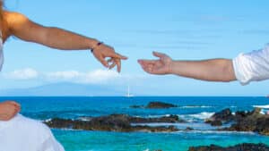 Two hands reaching out over a rocky ocean shore with a sailboat in the distance.