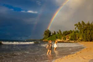 Two children play near the ocean under a vibrant rainbow on a cloudy day.